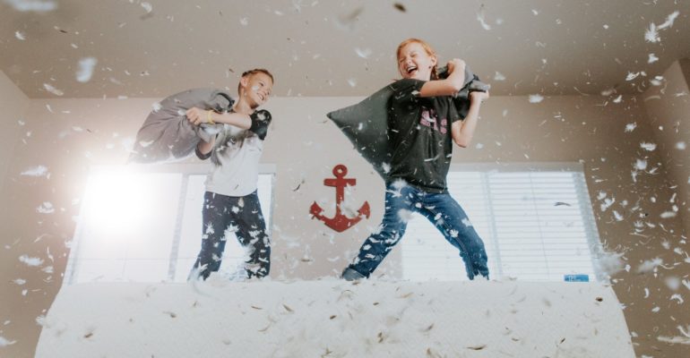 2 niños jugando sobre una cama con almohadas de plumas. Foto alegórica de la necesidad de contacto físico e interacción social que tienen los niños y adolescentes, producto del confinamiento y el distanciamiento social por la pandemia.